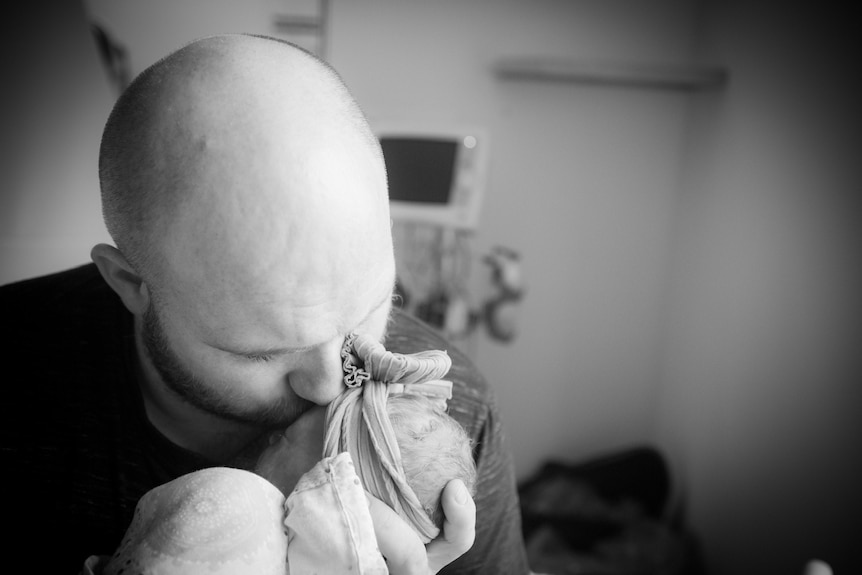 A man holds a newborn baby in his arms and kisses the side of her face.