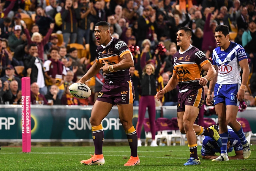 A male NRL player holds the ball in his right hand as he looks to the crowd while celebrating a try.