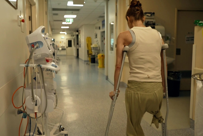 A woman with one leg uses crutches to walk down a hospital corridor