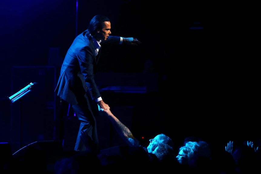 Nick Cave holds the hand of an audience member as he sings on stage in Sydney