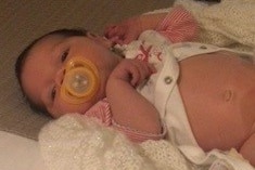 A newborn baby with a yellow dummy lays on a bed 