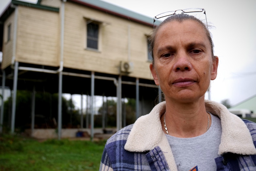 A woman in a flannelette shirt with glasses and a house on stilts in the background