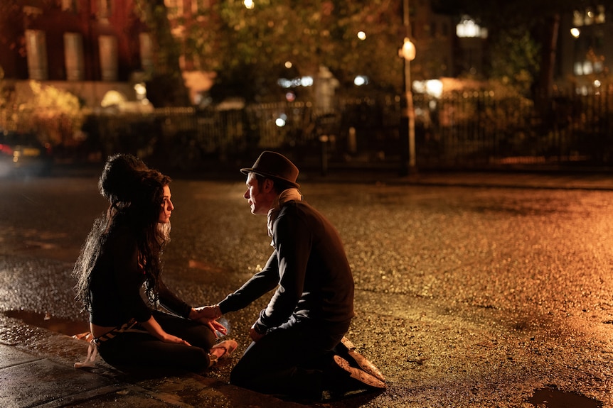 Amy sits on a curb as Blake leans over to her at nighttime, as they have a deep conversation.