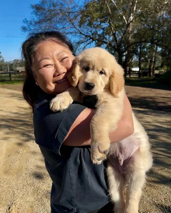 Nagi, standing in a field, holds up the golden puppy, smiling.