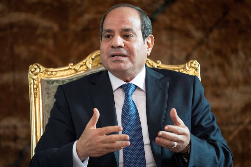 Abdel Fattah El-Sisi wearing a suit and blue tie sits in an ornate chair with golden frame