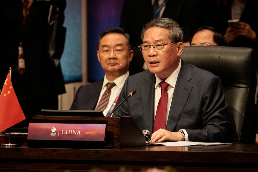 Li Qiang, wearing a suit with red tie, sits at a desk behind a name plate that says CHINA and a microphone