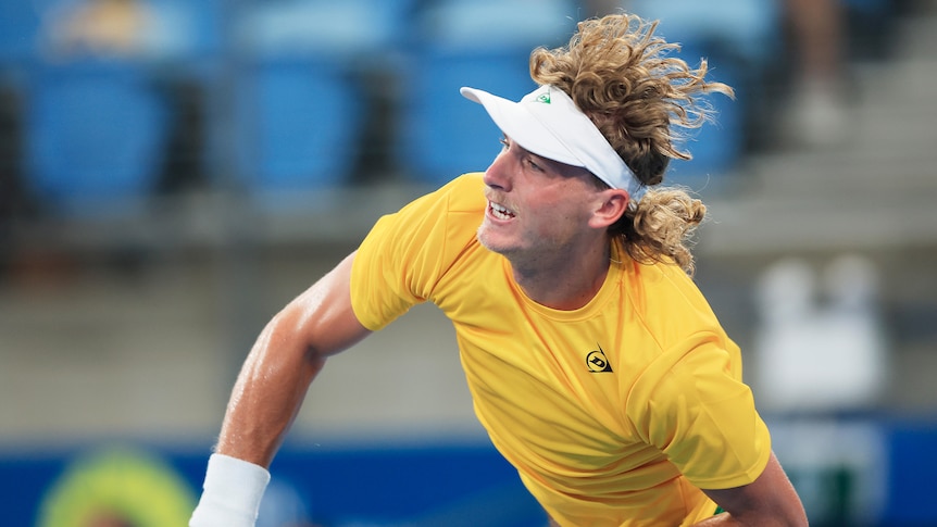 Max Purcell, wearing a gold shirt and white visor while representing Australia, serves during an ATP Cup tennis match.
