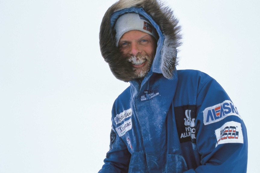 A man in a blue snow-jacket with a fur-lined hood smiles for a photo. His beard is covered with ice.