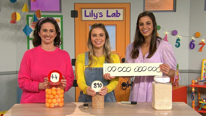 Lily Serna, Rachael and Emma on the Play School set all holding up different numbers, with the sign "Lily's Lab" on the wall.