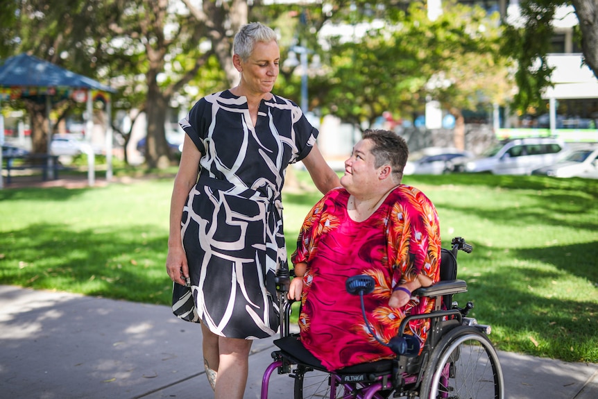 A woman in a black dress stands next to a woman wearing red in a wheelchair who has had her legs and hands amputated.