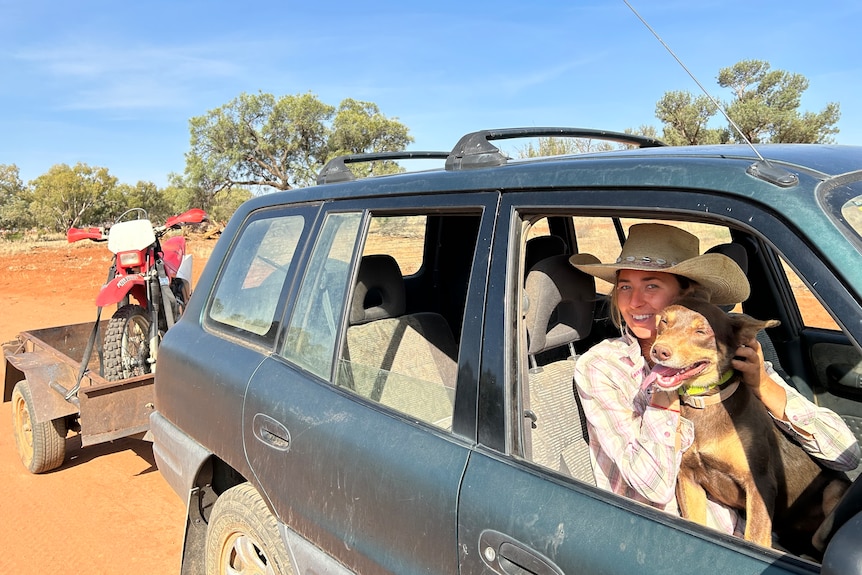 Girl in green car with her kelpie work dog looking out the window, and a trailer with a motorbike on the back 