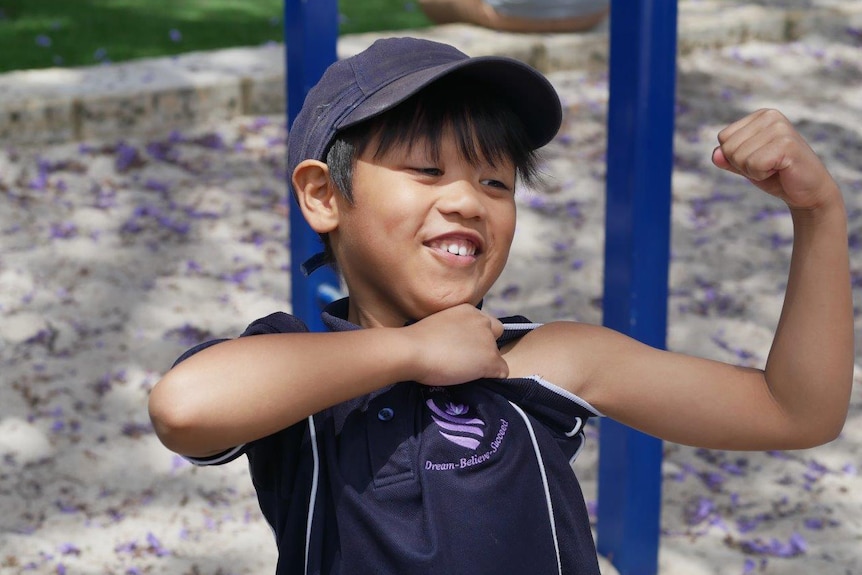 A young boy wearing his school uniform and a blue cap flexes his bicep in the playground.
