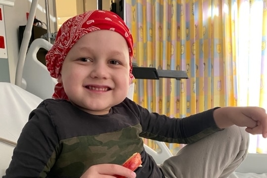 A young boy smiles wearing a hard scarf eating fruit.