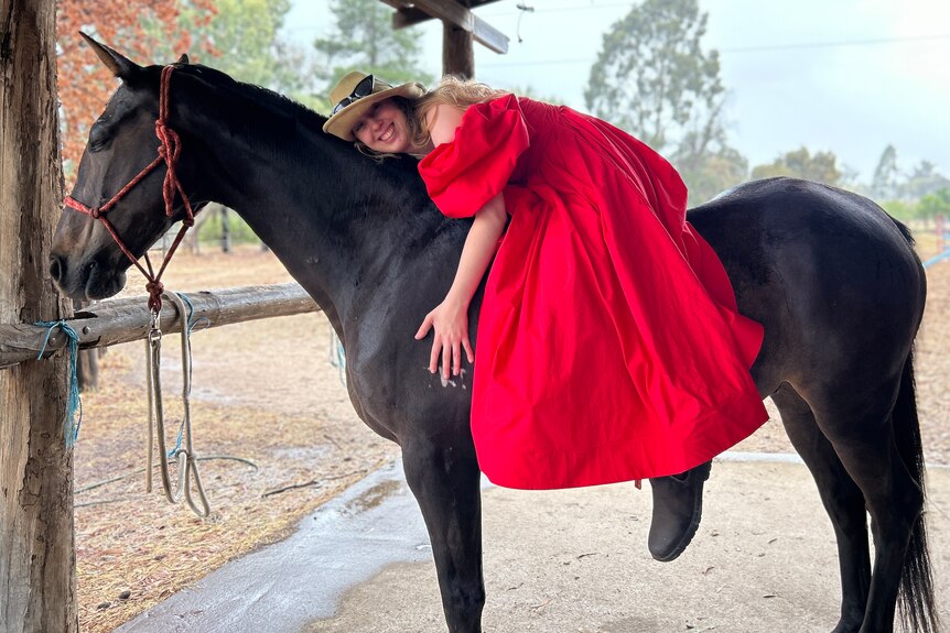 A young woman wearing a red gown, sitting on a horse resting her head on its neck.