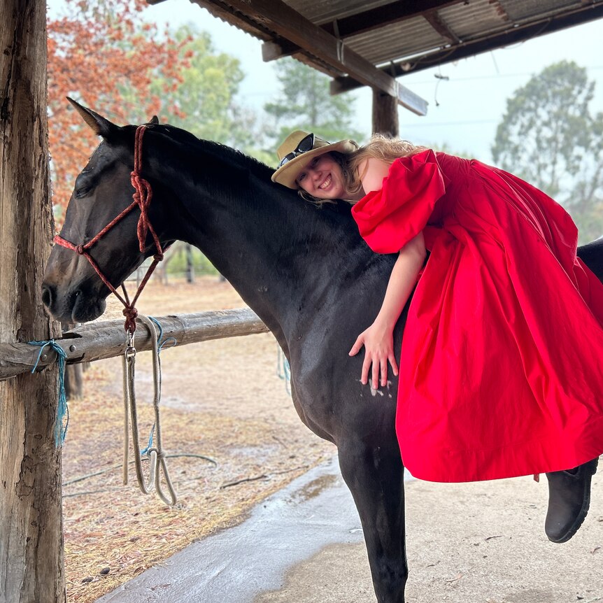 A young woman wearing a red gown, sitting on a horse resting her head on its neck.