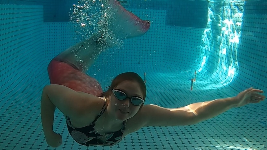 Woman with mermaid tail and swimming goggles swims underwater in pool