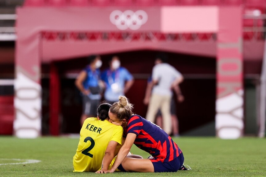 A soccer player in a yellow jersey sitting on the pitch being consoled by a player from the other team