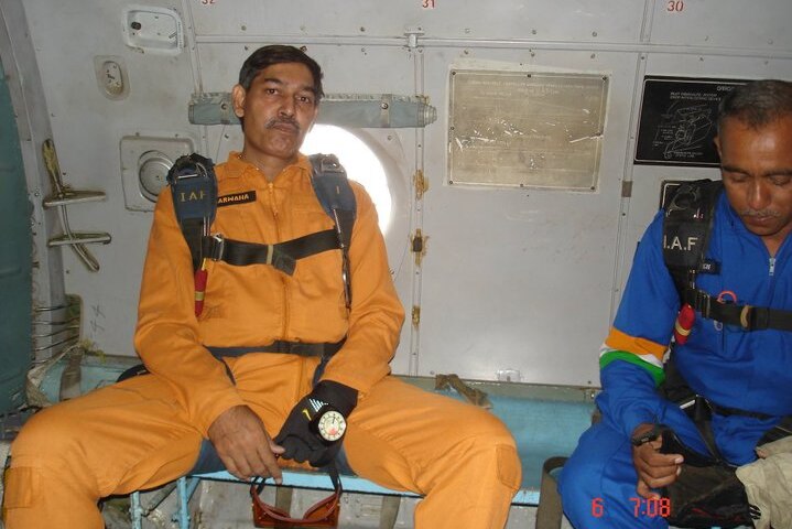 Arun Marwaha in an orange jumpsuit seemingly inside a plane, sits on a bench and looks at the camera with a serious expression