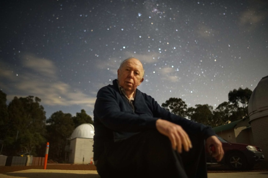 An elderly man sits in front of an observatory under a starry sky.