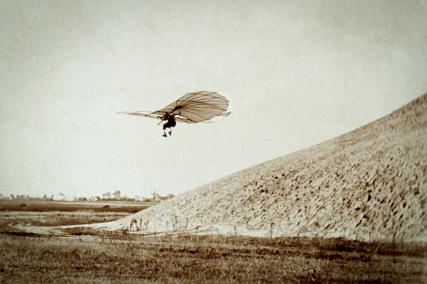 Man flying a very early glider just above the ground and a small hill.