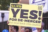 A person holds a sign that reads 'Yes for de-merger, 62.5 per cent voted'