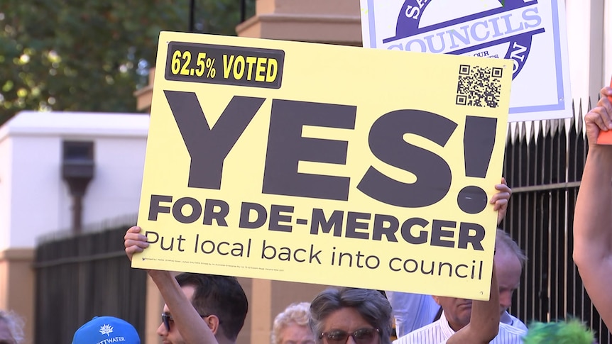 A person holds a sign that reads 'Yes for de-merger, 62.5 per cent voted'
