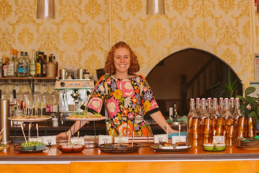 A woman with short, curly ,red hair in a floral dress posing behind an orange tiled bar.