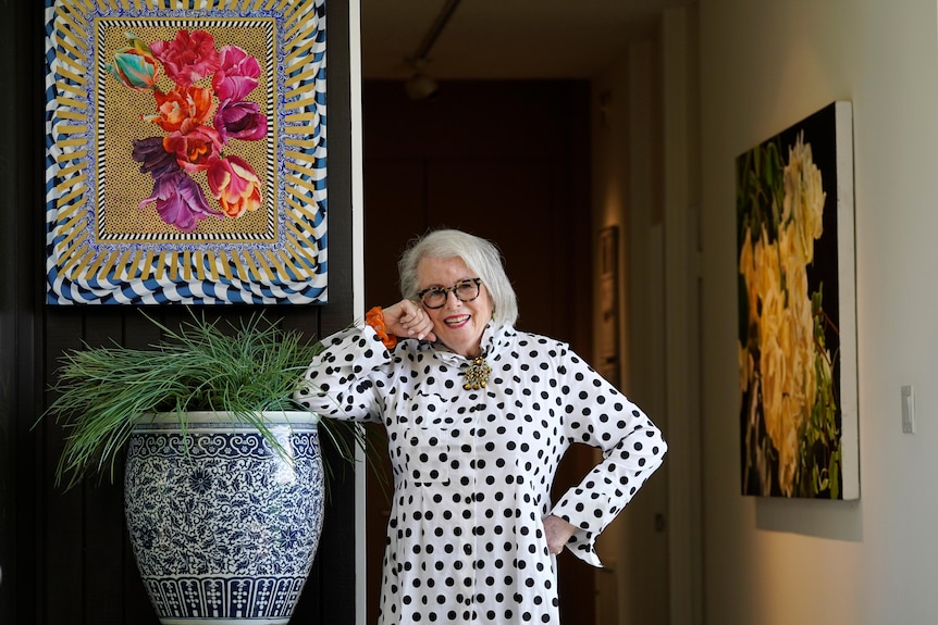 An older woman wearing a black and white polka dot dress leaning on a plant pot 