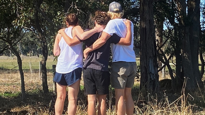 Three people stand with their arms around each other in front of some trees.