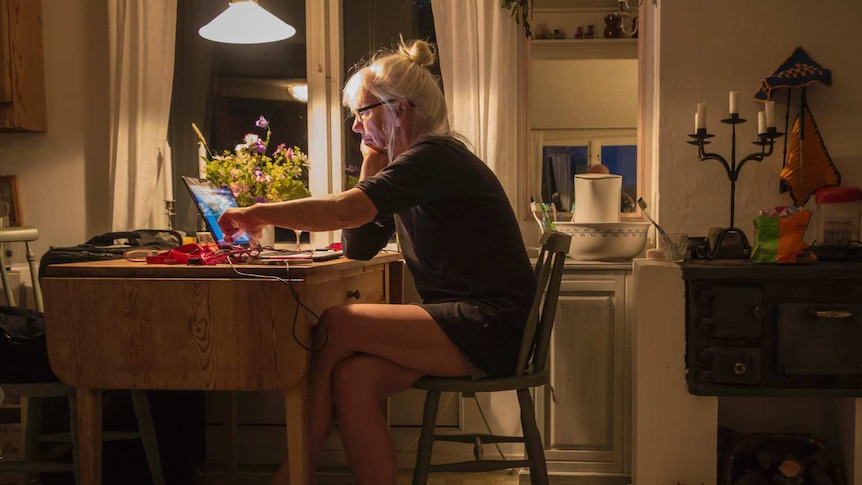 An elderly woman with grey hair sits at a table staring at a computer screen.