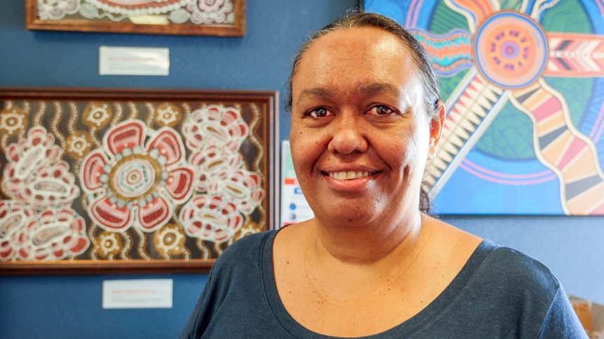 An Indigenous woman standing in front of Aboriginal artwork wearing a dark blue shirt