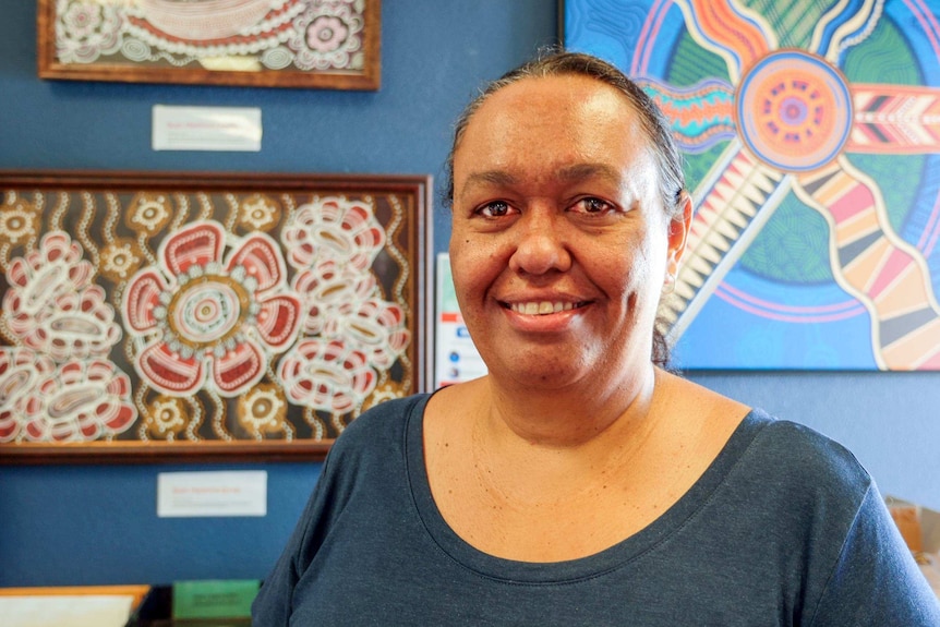 An Indigenous woman standing in front of Aboriginal artwork wearing a dark blue shirt