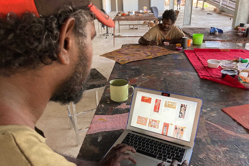 Shaun Namarnyilk accessing the Injalak.com shop with Amos Nganjmirra in the background. There is painting going on around them.