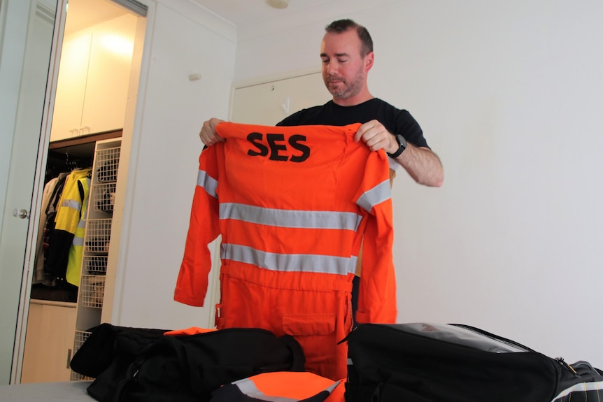 A wide shot of a man standing in a bedroom packing away his emergency services protective clothing.