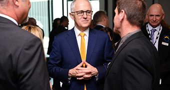 A mid-shot of a smiling Malcolm Turnbull, wearing a blue suit and yellow tie, listening to a man talking to him.