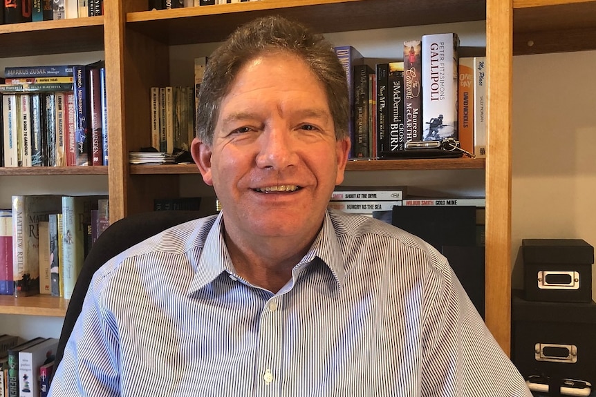 Michael Gray smiles, dressed in a blue shirt as he sits in front of a full bookshelf.