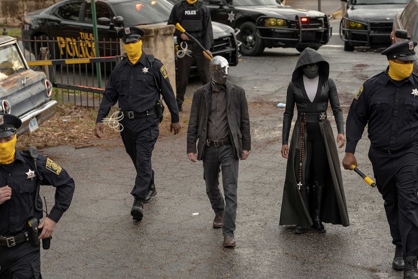 A scene from the HBO series Watchmen with two superheroes Looking Glass and Sister Night and police in masks