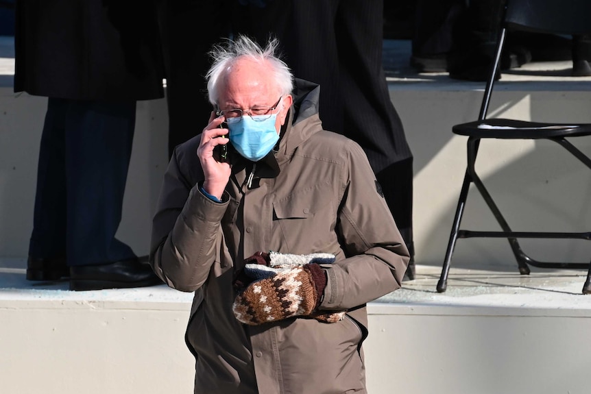 Bernie Sanders in a brown parka and blue face mask speaking on the phone, while clutching cosy mittens