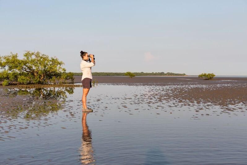 A woman stands on mudflats looking through binoculars.