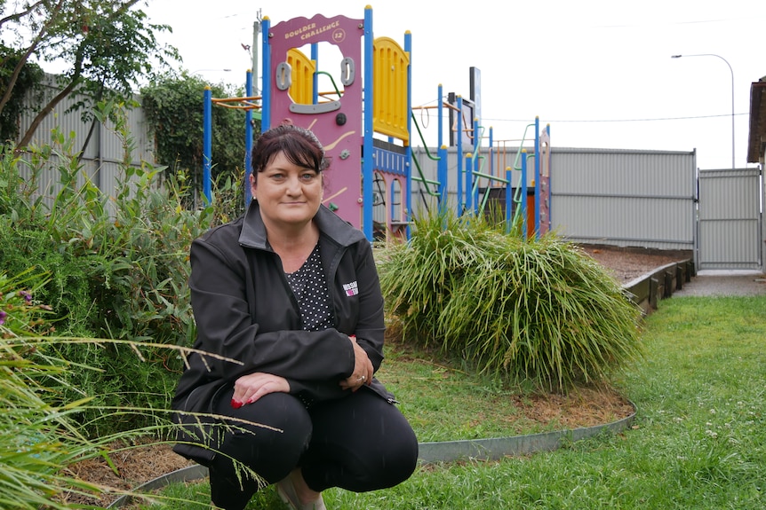 A woman in a black jacket kneeling down in a garden by a playground.