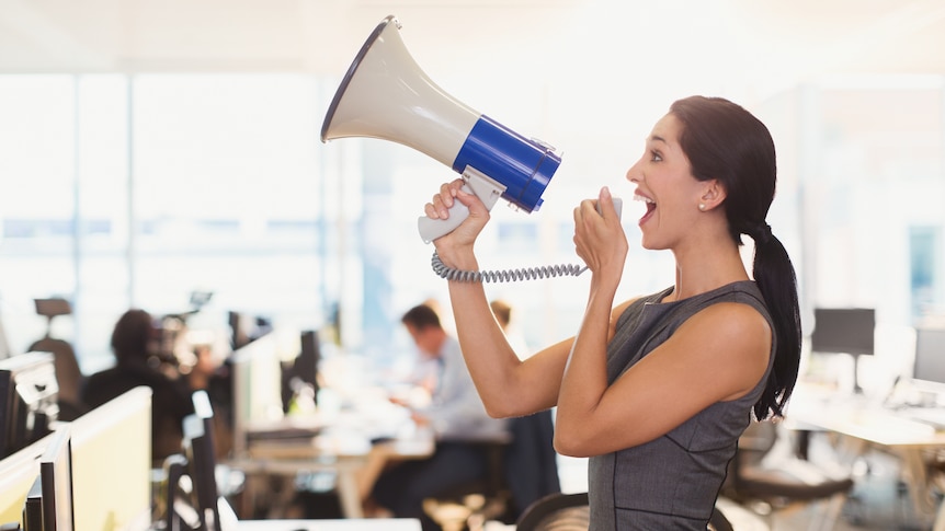 A woman, dressed in a grey dress, uses a megaphone in the office.