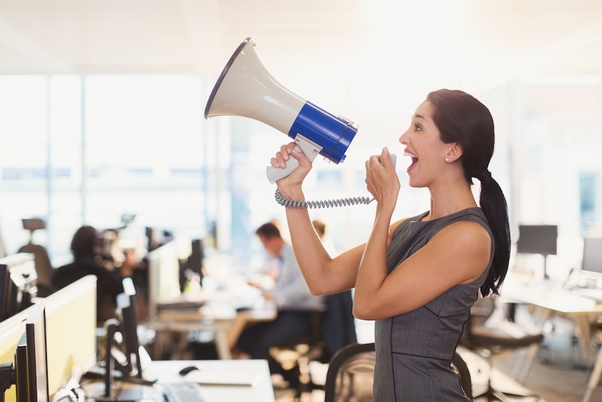 A woman, dressed in a grey dress, uses a megaphone in the office.
