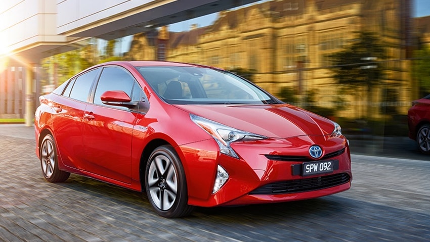 A red Toyota Prius car drives on a cobblestone street next to a reflective glass window.