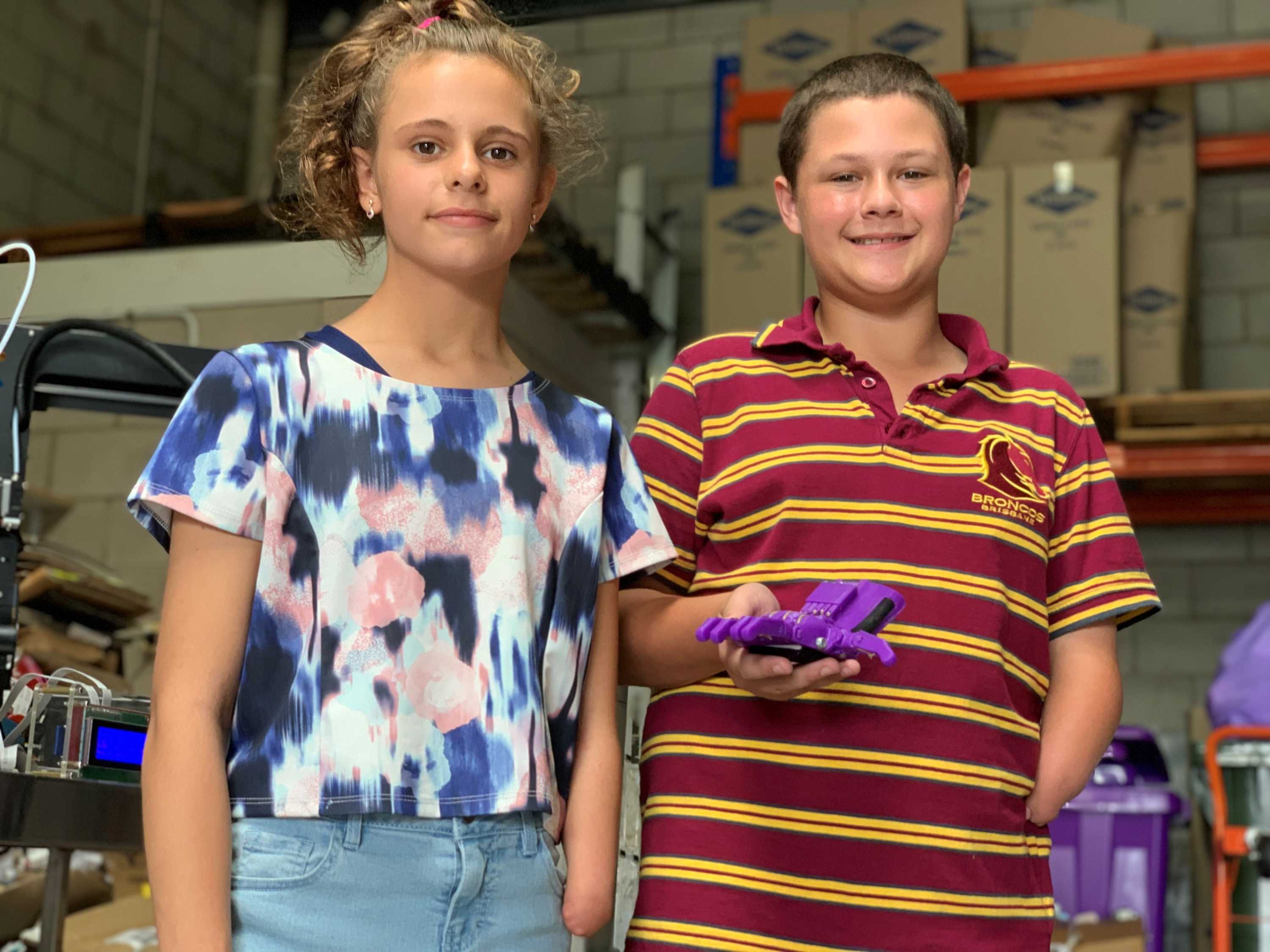 Una niña y un niño parados en un almacén, el niño sostiene una mano protésica de plástico violeta impresa en 3D.