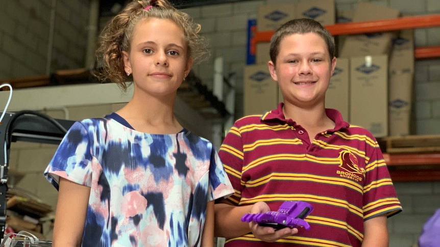 A girl and a boy stand in a warehouse, the boy is holding a 3-D printed purple plastic prosthetic hand.