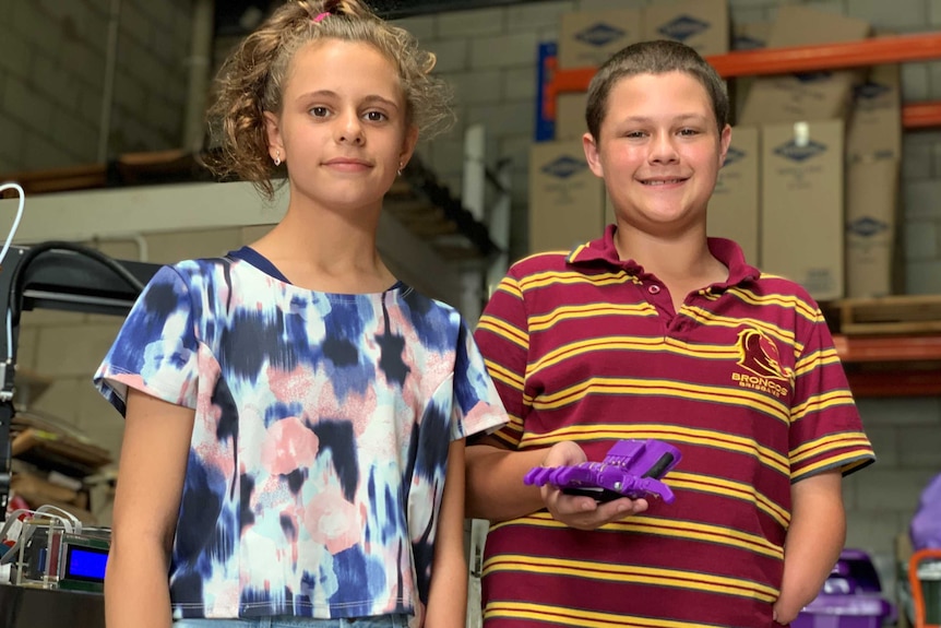 A girl and a boy stand in a warehouse, the boy is holding a 3-D printed purple plastic prosthetic hand.