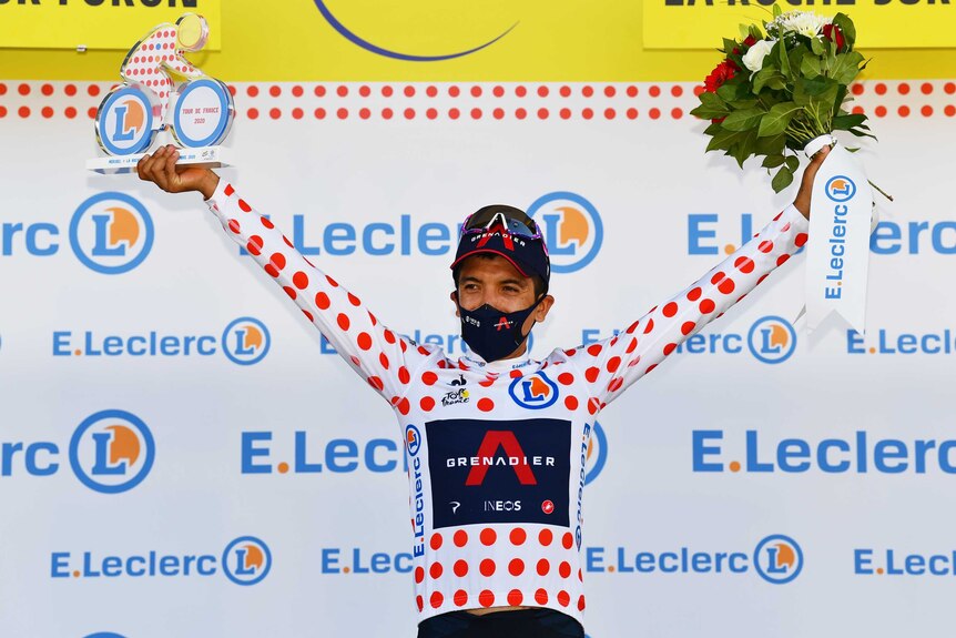 Richard Carapaz stands arms aloft wearing the polka-dot jersey on a Tour de France dais.