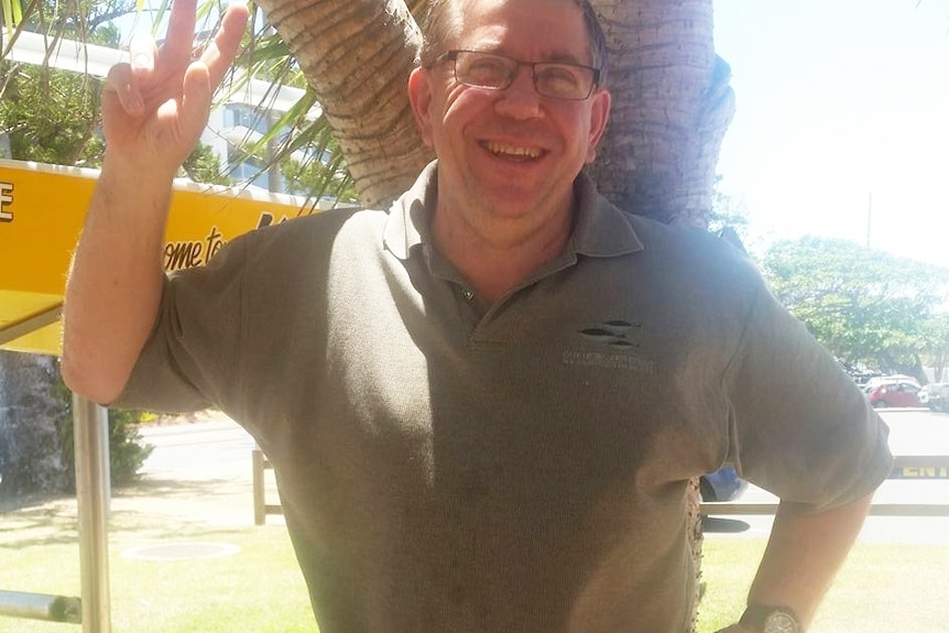 Bruce Saunders smiles as he makes a peace sign with his right hand while standing under a tree, location unknown.