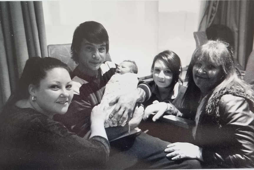 A black and white photo of a young man holding a baby while two women and a young girl crouch next to him.