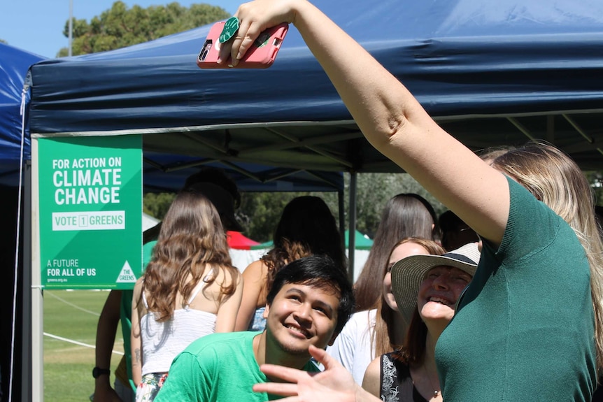 Two young Greens members take a selfie in front of the Greens tent which includes a sign saying "For action on Climate Change".